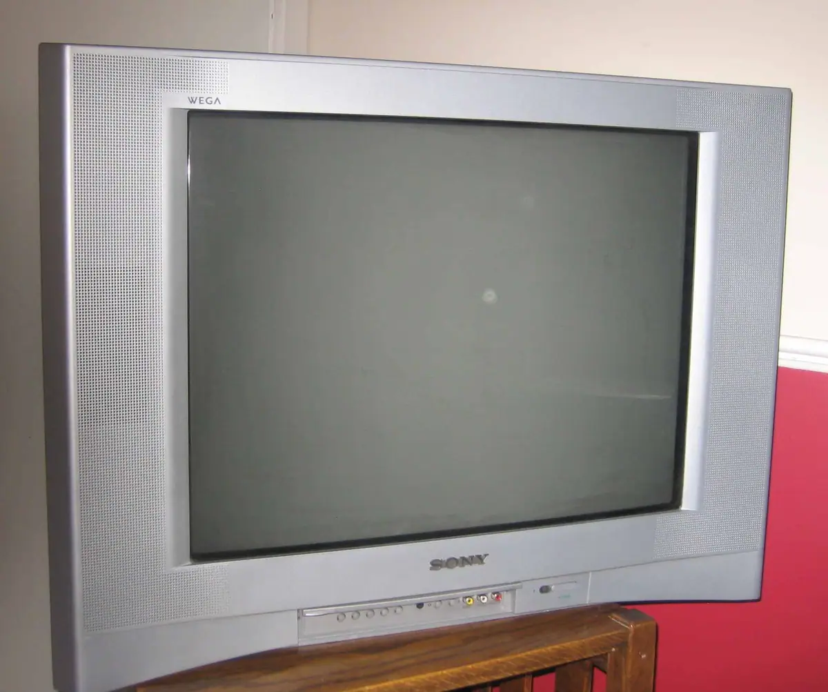A silver CRT television sits on a wooden stand against an interior wall.