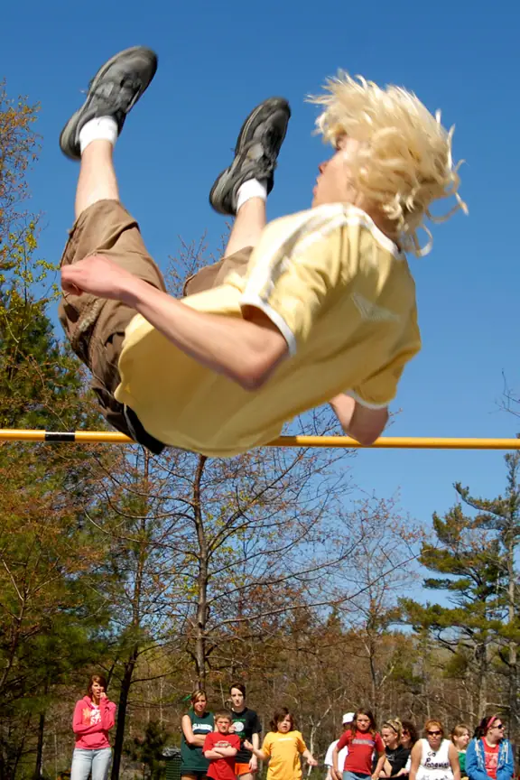 A person clears a yellow bar while spectators watch in a wooded outdoor setting.
