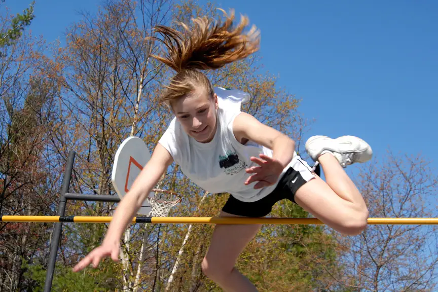 A young athlete clears a yellow bar outdoors with a basketball hoop and trees behind her.